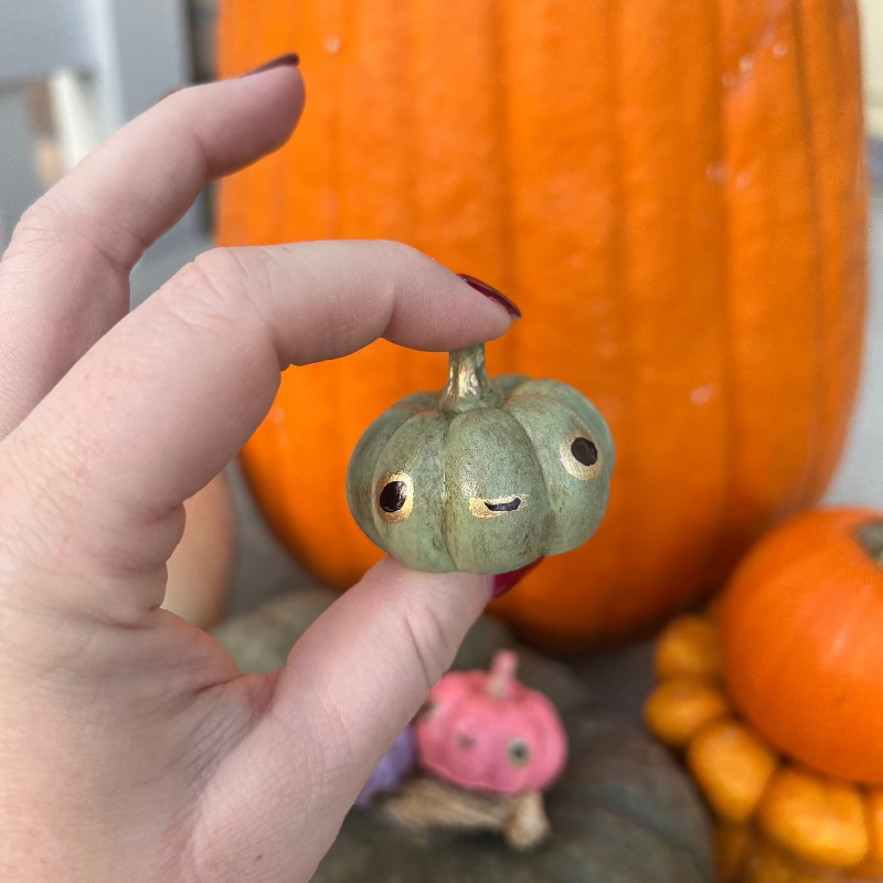 Hand holding a small decorative pumpkin with a face in front of larger pumpkins.