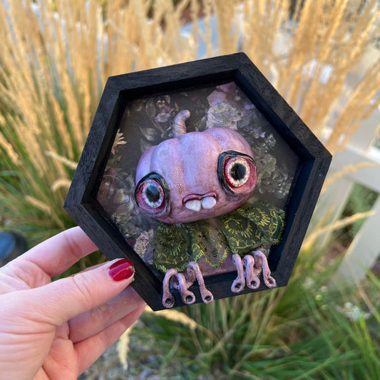 Hexagonal framed artwork of a pumpkin creature held by a hand outdoors.