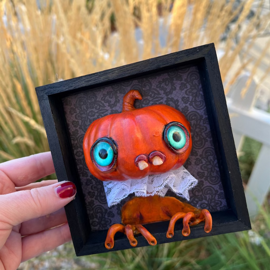 Hand holding a small pumpkin sculpture with a face in a black frame against a natural background.