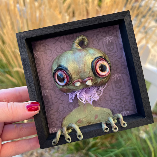 Small framed artwork of a green pumpkin creature with large eyes held by a hand against a natural background.