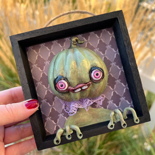 Small framed artwork of a creature with a pumpkin head held by a hand against a natural background.