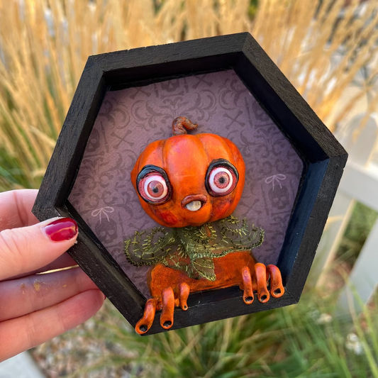 Small pumpkin creature in a black hexagonal frame held by a hand with red nail polish, against a natural background.