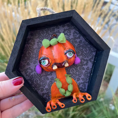 Hexagonal black frame with a decorative pumpkin monster figure held by a hand.