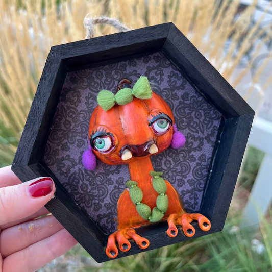 Hexagonal black frame with a decorative pumpkin monster figure held by a hand.