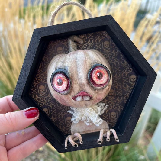 Hexagonal framed artwork of a creature with large eyes held by a hand outdoors.