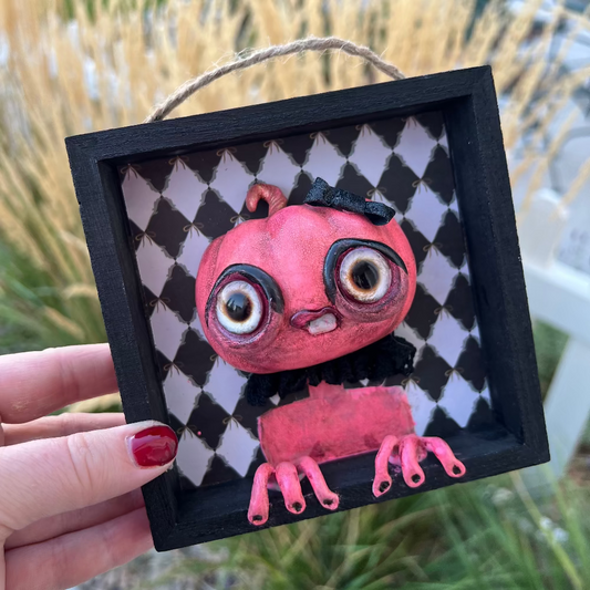 Small pink creature art piece in a black frame held outdoors.