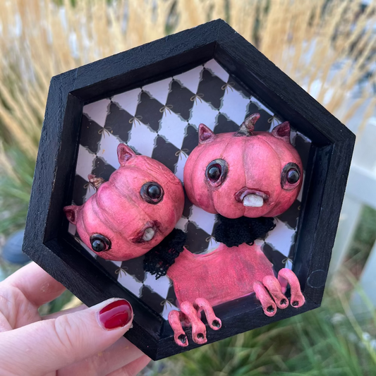 Hexagonal black frame with pink cartoonish creatures on a checkered background, held by a hand.