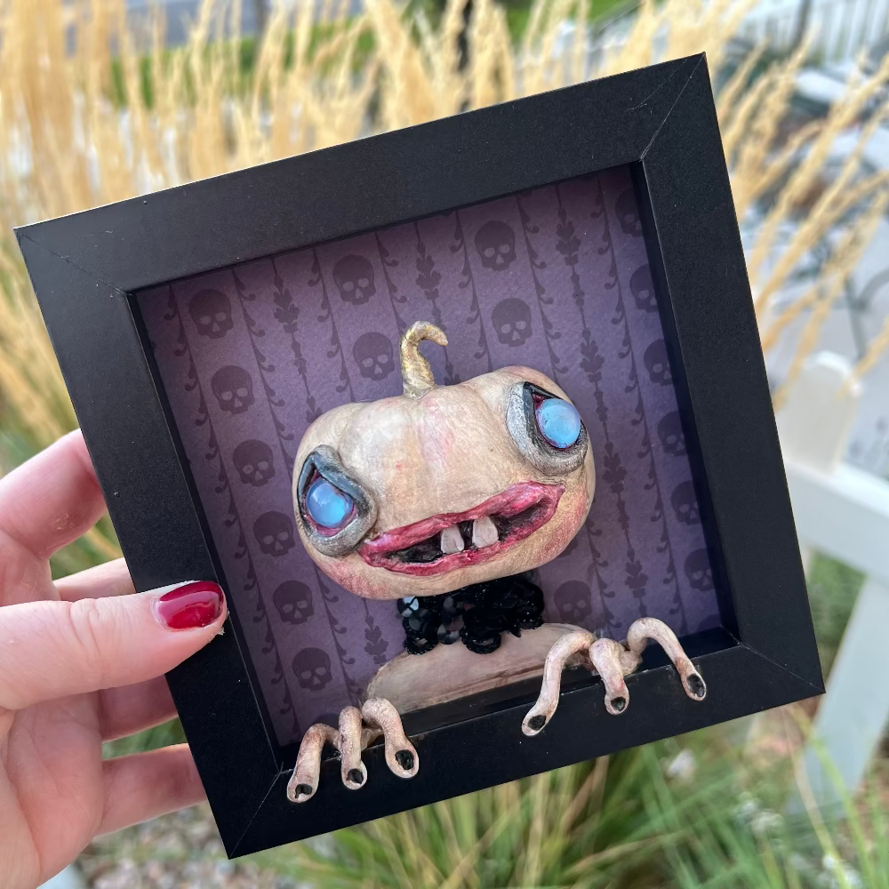 Framed artwork of a creature with large eyes and a black frame held by a hand.