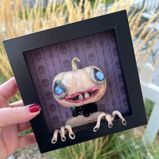 Framed artwork of a creature with large eyes and a black frame held by a hand.