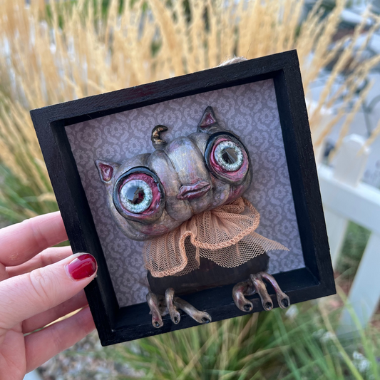 Small framed artwork of a creature cat kitty  pumpkin with large eyes held by a hand outdoors.