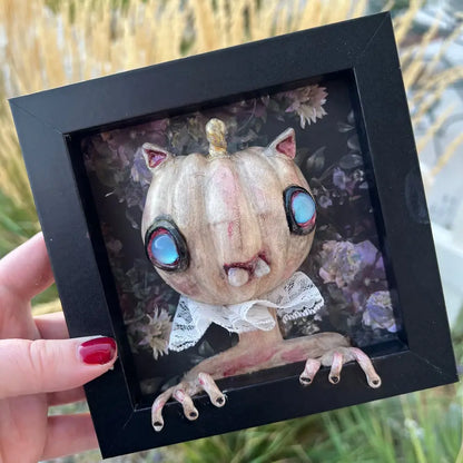 Framed artwork of a creature with pumpkin head and large eyes held by a hand.