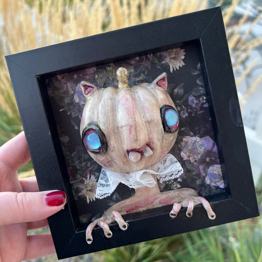 Framed artwork of a creature with pumpkin head and large eyes held by a hand.