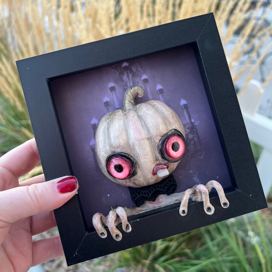 Framed artwork of a pumpkin with googly eyes held by a hand outdoors.
