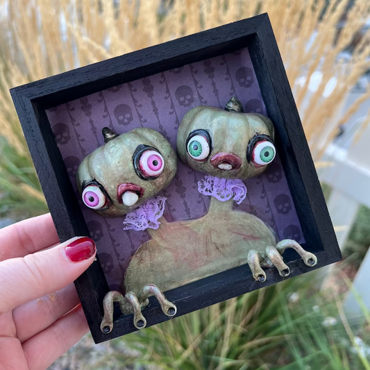 Framed artwork of two green alien-like creatures with googly eyes held by a hand.