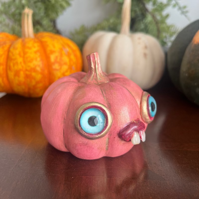 Pink pumpkin with monster face decorations on a reflective surface with pumpkins in the background.