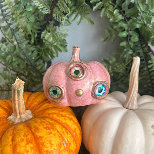 Decorative pumpkins with a pink one featuring googly eyes against a green leafy background