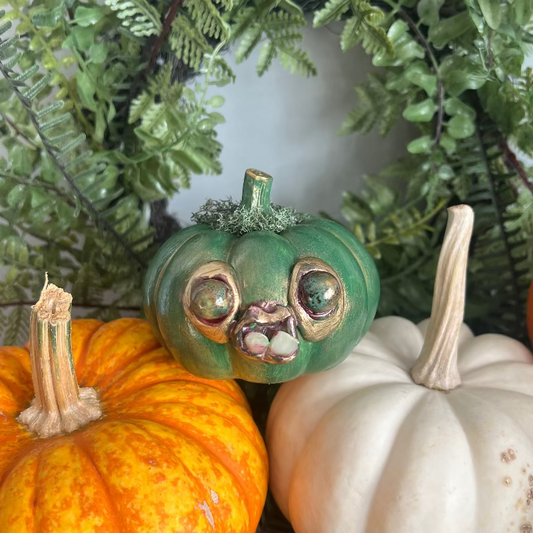 Decorative pumpkins with a green one featuring eyes and a mouth, surrounded by greenery.