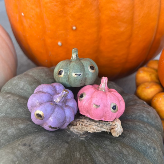 Three small painted pumpkins in purple, green, and pink on a larger pumpkin with a blurred background.