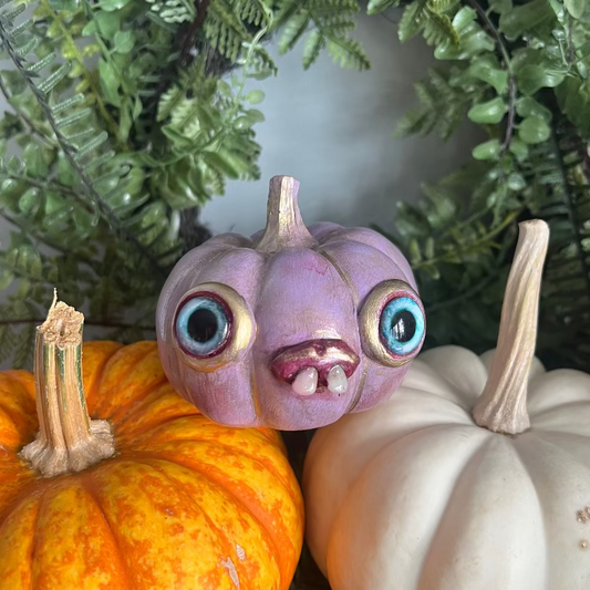 Purple pumpkin figurine with large eyes among real pumpkins and greenery