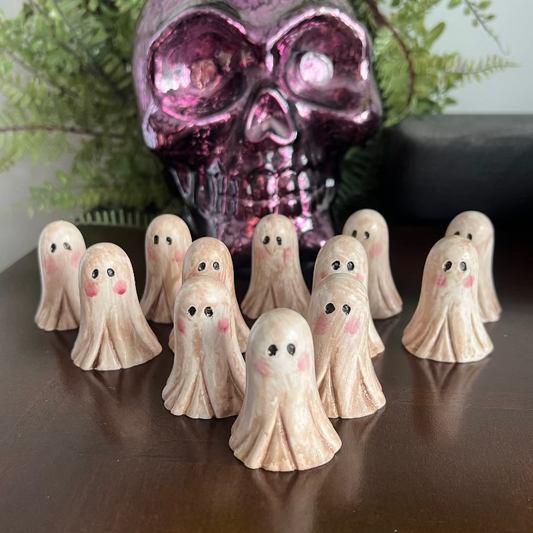 Small ghost figurines with pink cheeks on a reflective surface with a metallic skull in the background.