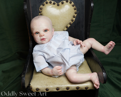 A realistic reborn baby doll with pale skin and blue eyes, wearing a light blue dress, reclining on a vintage-style armchair with a heart-shaped backrest against a dark green fabric backdrop.