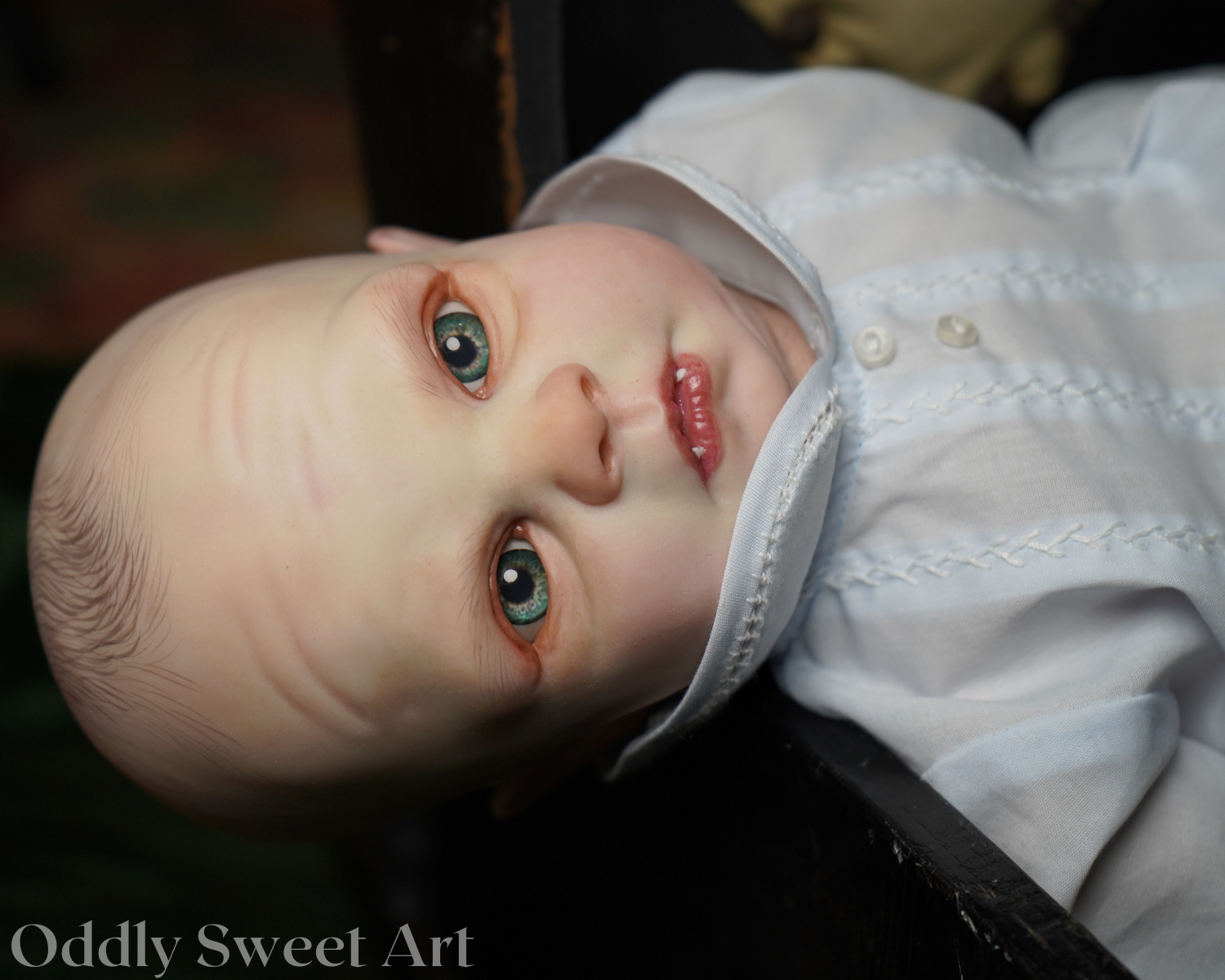 A close-up photograph of a realistic baby doll with large blue-green eyes, pale skin, and pink lips, wearing a white embroidered bonnet and white dress. The doll is lying on a dark surface, and a watermark reading 'Oddly Sweet Art' is visible in the lower-left corner.