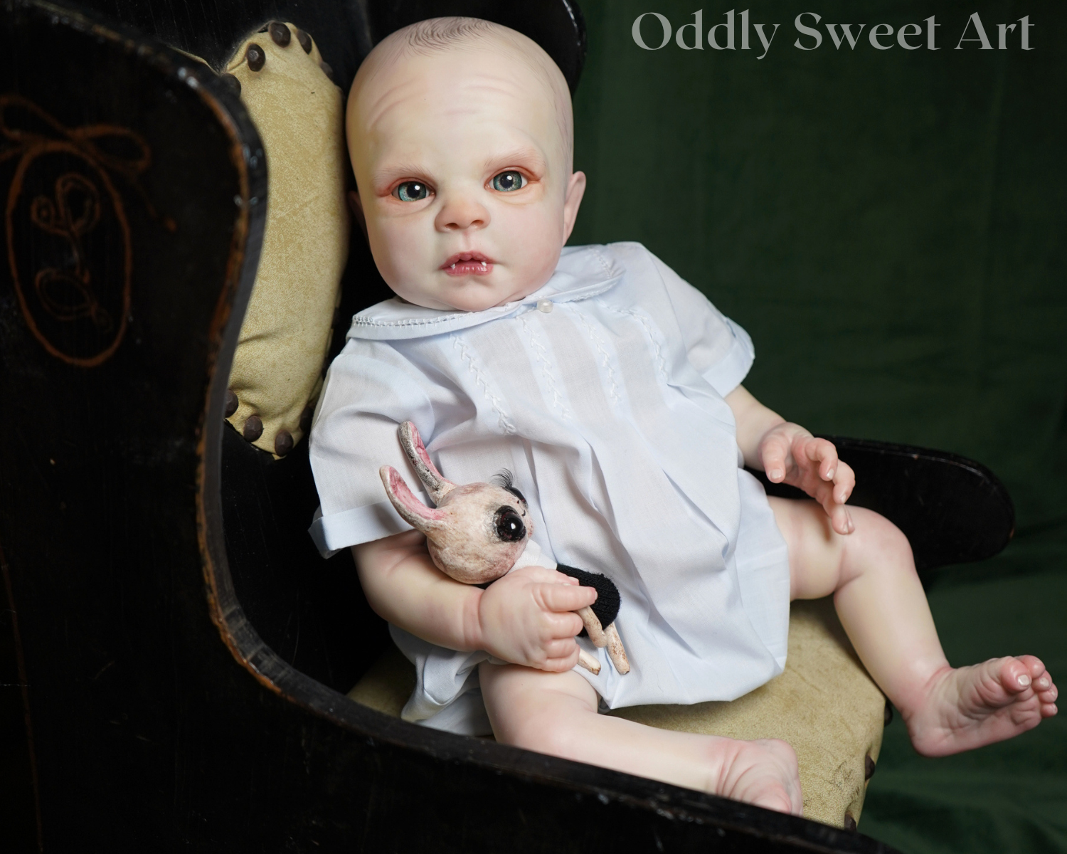 A realistic reborn baby doll with pale skin and blue eyes, wearing a white dress and holding a small stuffed rabbit, seated in a dark wooden chair against a dark green backdrop. The text 'Oddly Sweet Art' is visible in the top right corner.