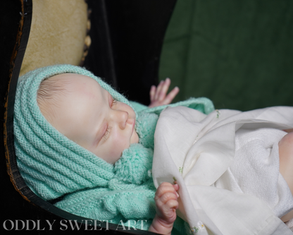 A close-up photograph of a sleeping newborn baby wrapped in a mint green knit bonnet and swaddle, resting on a dark wooden chair against a dark green background. The text 'ODDLY SWEET ART' is visible as a watermark at the bottom left.