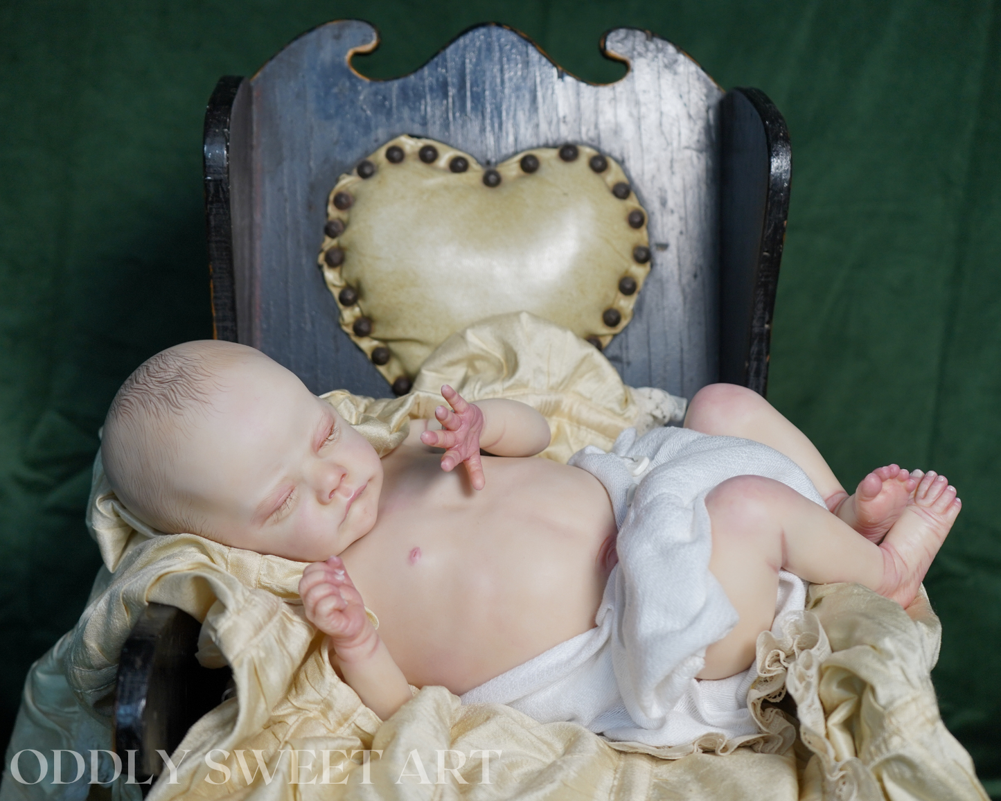 A realistic newborn baby doll or prop baby lying on cream fabric draped over a dark wooden chair with a heart-shaped tufted backrest, set against a dark green backdrop. A watermark reading 'ODDLY SWEET ART' is visible in the lower left corner.