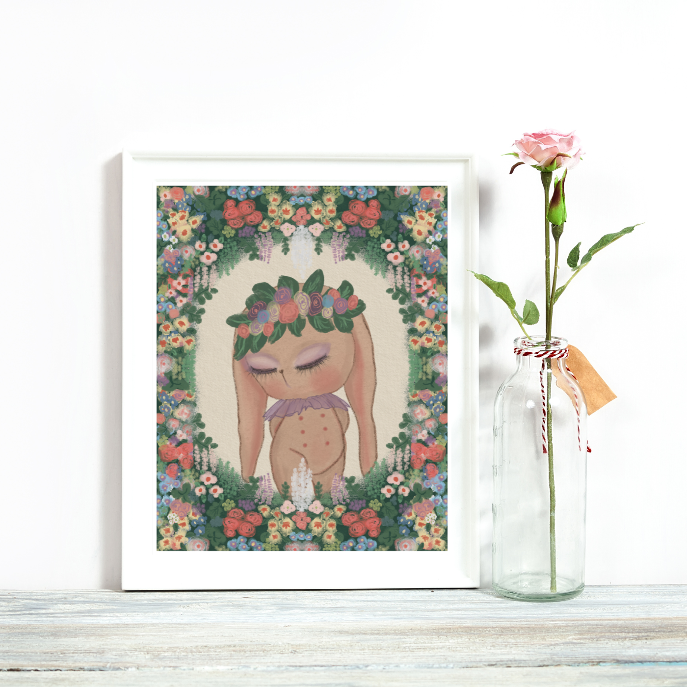 Framed artwork of a woman with flowers and a vase with a single pink rose on a white surface.