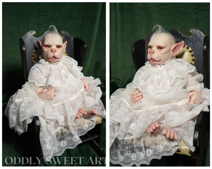 A side-by-side comparison of a realistic reborn doll with pointed ears, dressed in a white lace gown, seated in a dark wooden chair against a dark green background. The bottom left corner features the text 'ODDLY SWEET ART'.