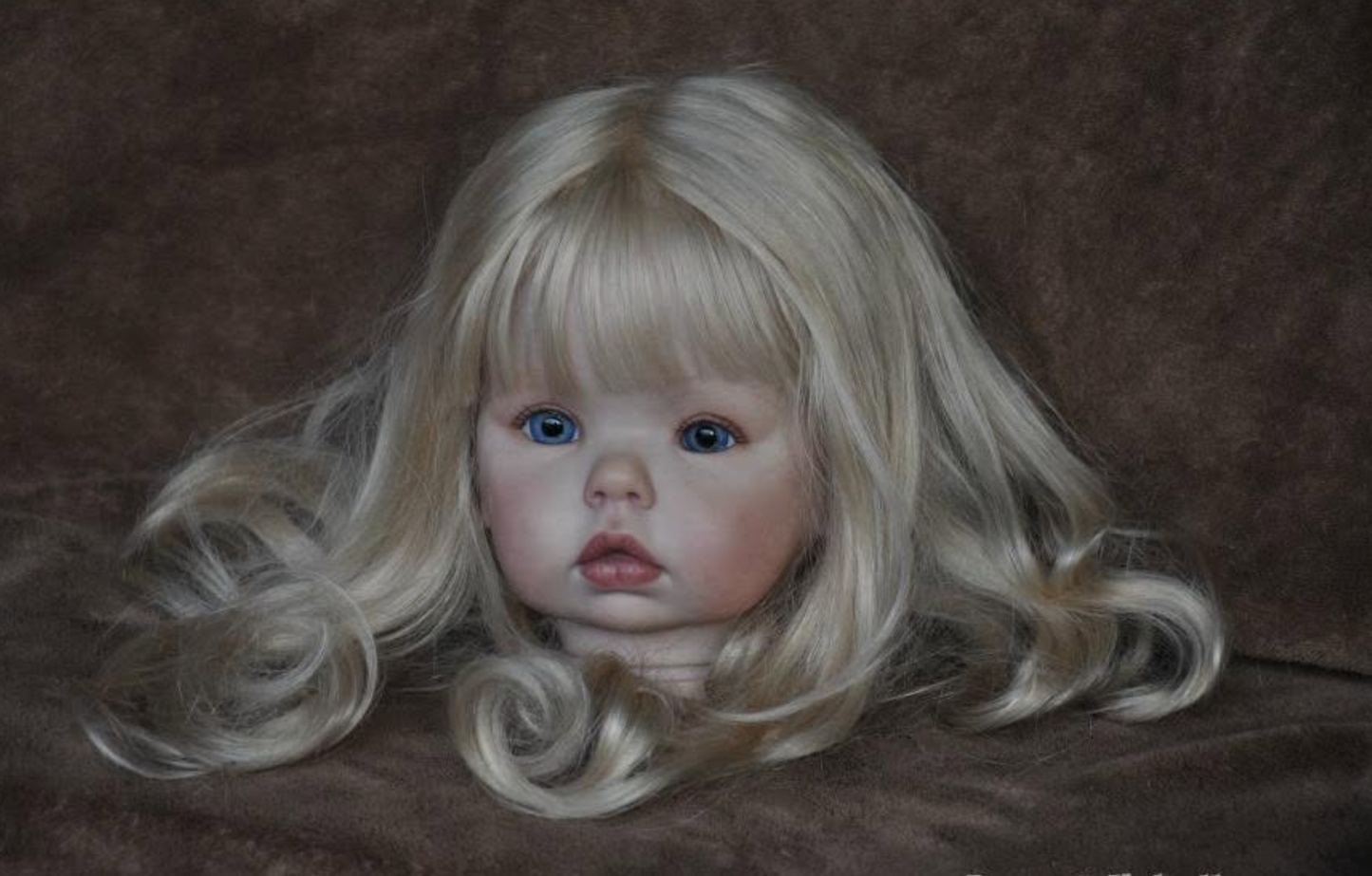 A realistic blonde doll head with long wavy hair, blue eyes, and a pale pink face, photographed against a dark brown textured background.