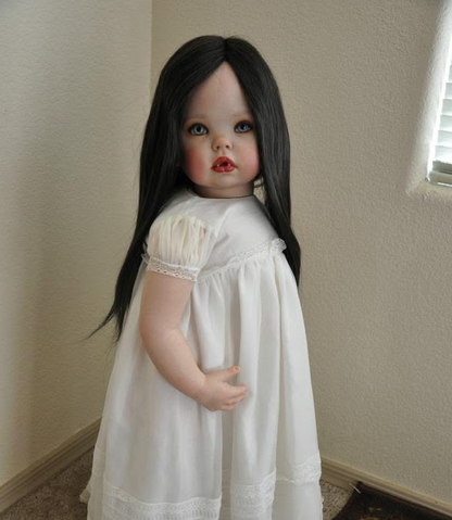 A realistic porcelain doll with long black hair, blue eyes, and pale skin, wearing a white short-sleeve nightgown with lace trim, standing in a room corner.