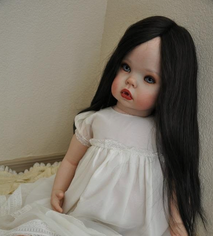A realistic porcelain doll with long black hair, blue eyes, and pale skin, wearing a white nightgown with lace trim, posed against a textured beige wall.