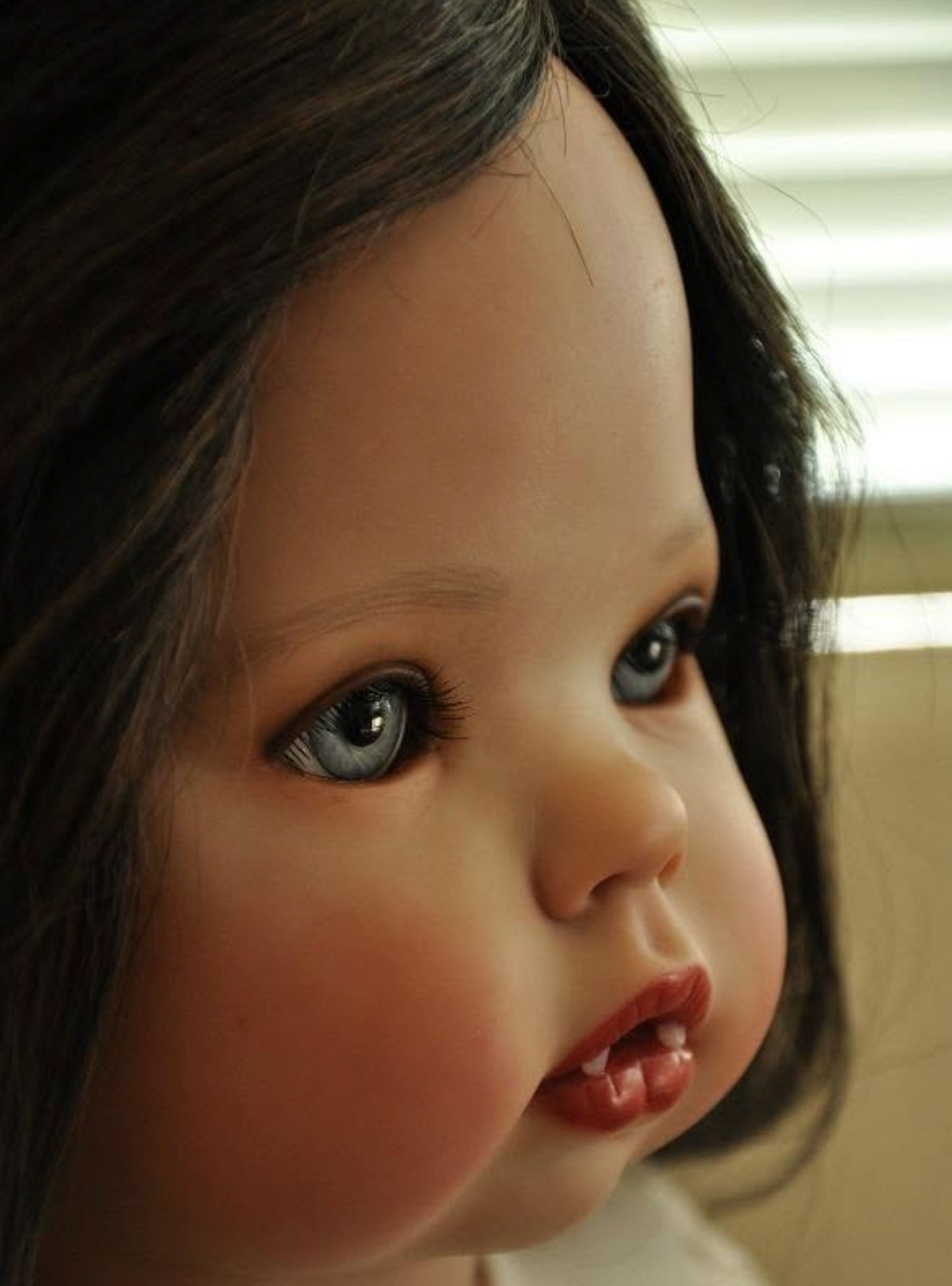 Close-up of a realistic doll or toy face with dark brown hair, large blue-gray eyes, rosy cheeks, and glossy red lips, shown against an indoor blurred background.