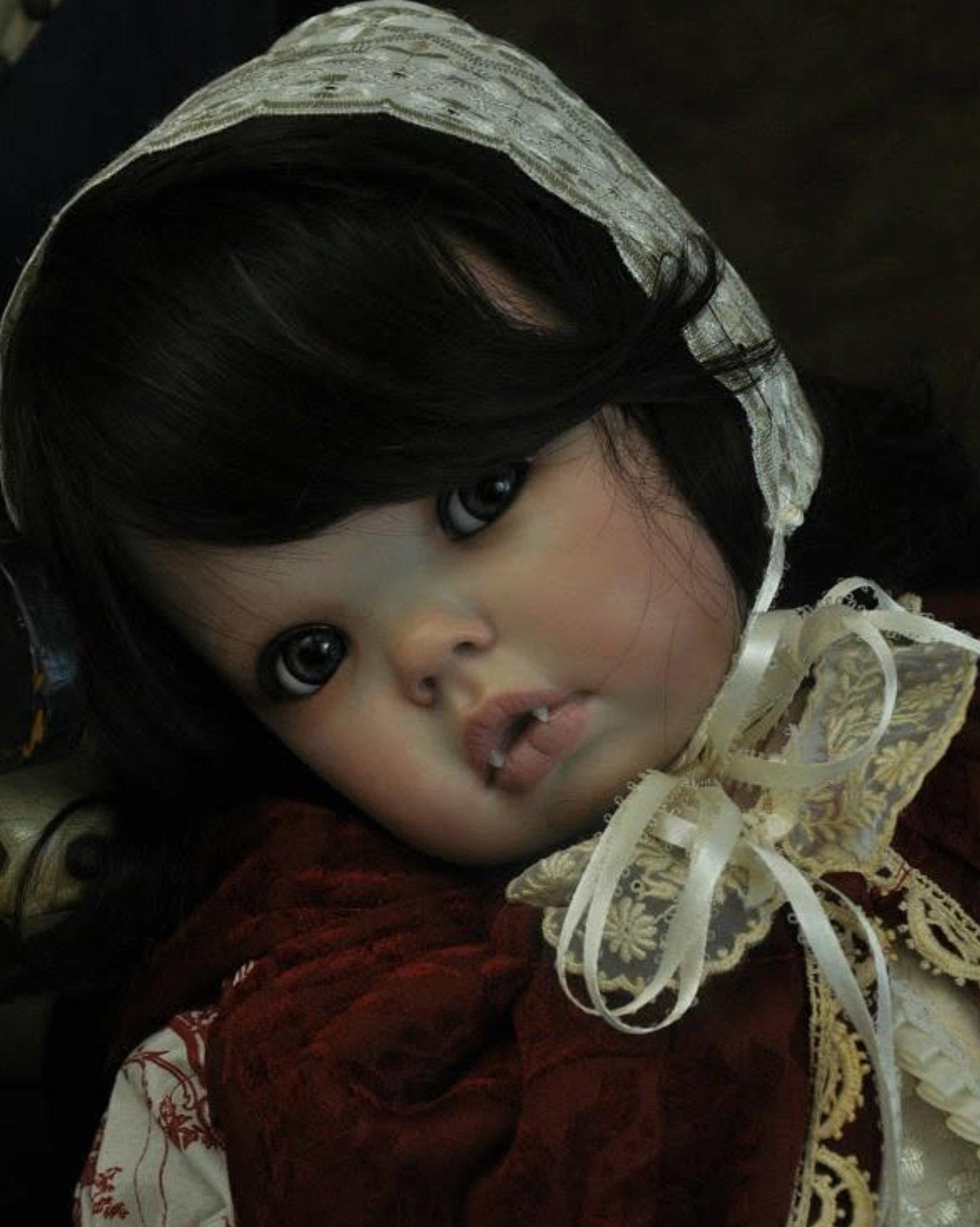 A close-up of a realistic porcelain doll with dark hair, large glossy eyes, and a cream lace bonnet and bow, wearing a deep red dress.