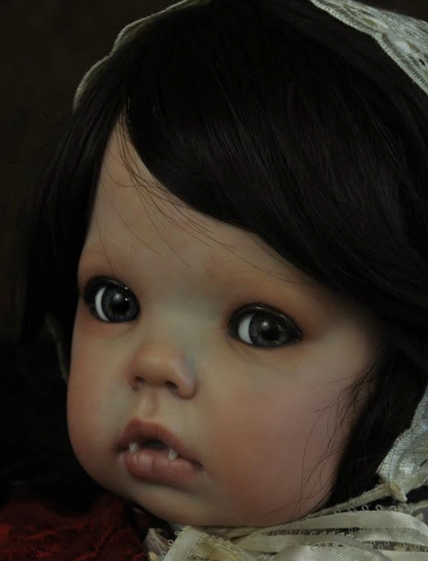 Close-up of a realistic baby doll with dark hair, large dark eyes, and a pale face, wearing a lace-trimmed bonnet and red clothing.