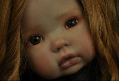 Close-up of a realistic doll or toy face with light skin, large dark eyes, pinkish lips, and strawberry-blonde hair against a dark background.