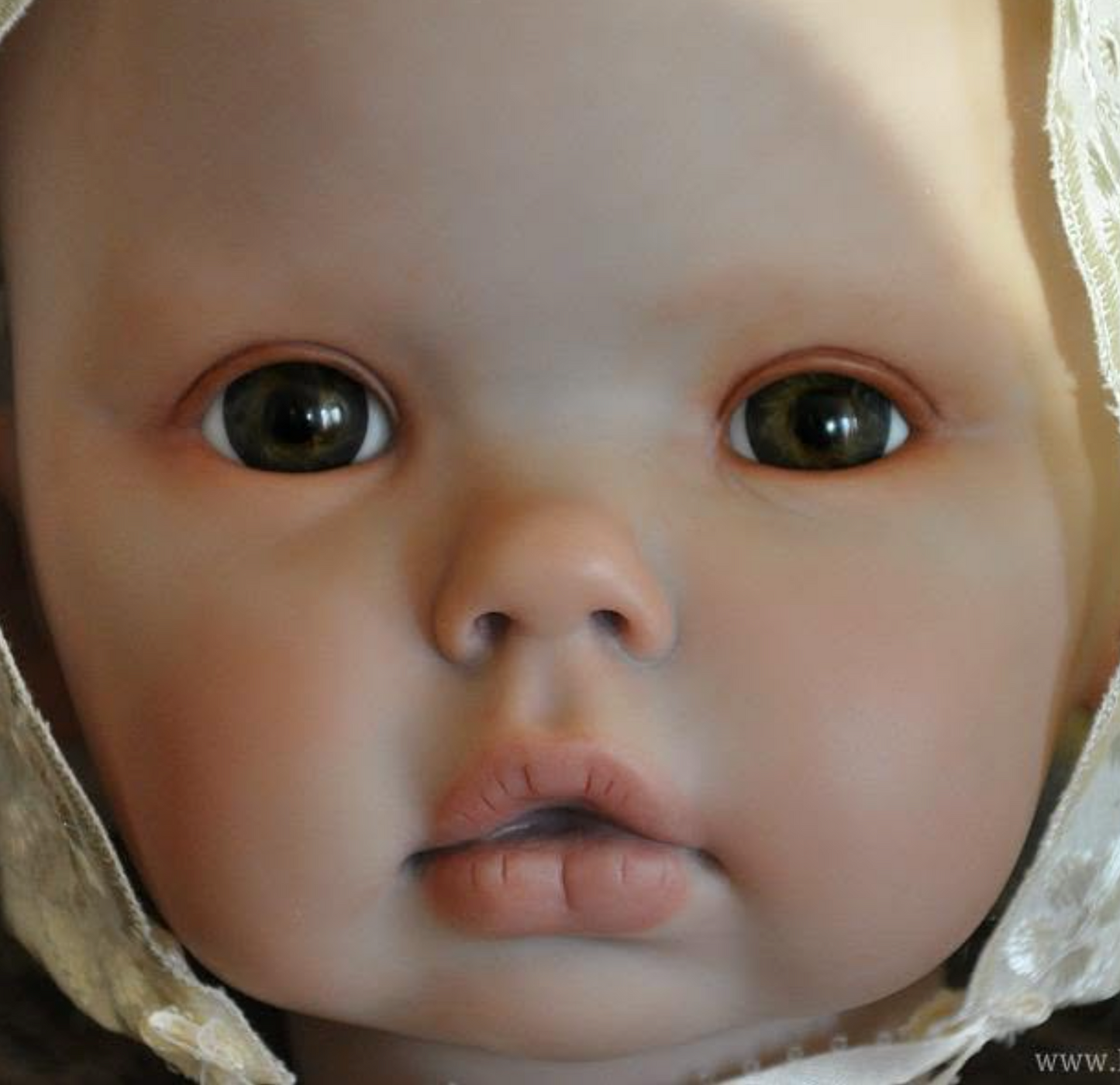 Close-up of a realistic baby doll or toy face wearing a cream lace bonnet, filling the frame.