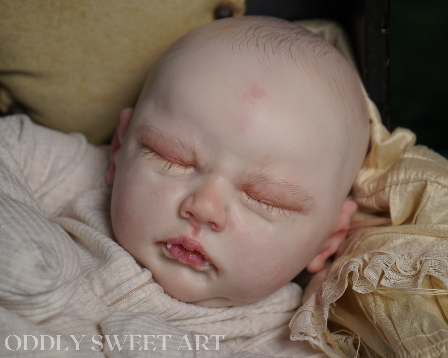 A close-up photograph of a realistic sleeping doll or prop baby wrapped in cream fabric, resting on a textured beige cushion. The watermark 'ODDLY SWEET ART' is visible in the bottom-left corner.
