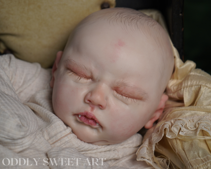 A close-up photograph of a realistic sleeping doll or prop baby wrapped in cream fabric, resting on a textured beige cushion. The watermark 'ODDLY SWEET ART' is visible in the bottom-left corner.